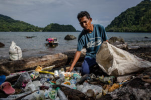 Ein Einwohner einer Südseeinsel sammelt Plastikmüll von einem Strand ein. Er schaut in die Kamera. Im Hintergrund sind Meer und bewaldete Berge zu sehen.