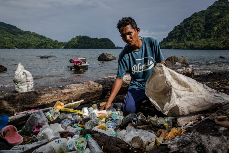 Ein Einwohner einer Südseeinsel sammelt Plastikmüll von einem Strand ein. Er schaut in die Kamera. Im Hintergrund sind Meer und bewaldete Berge zu sehen.