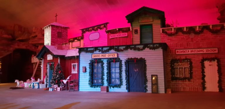 Ein Bühnenbild von einer Hausfassade im Western-Stil, weihnachtlich geschmückt und mit Schildern wie "Santas Post Office" und "Reindeer Breeding Station"
